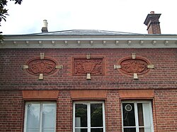 colour photo of brick wall with three picture inlaid in the brick, a central shield bearing three fleur de lis flanked by two faces, all three elaborately framed. Also windows below and roof above.