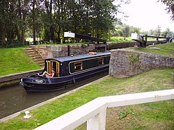 Narrow boat (named Toad) emerging from lock with black gates and white ends of the gate arms. Around the lock is a grassy area.