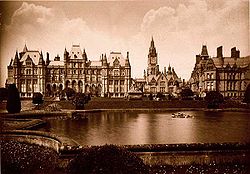 A sepia photograph with an artificial lake in the foreground, behind which are Gothic-style buildings, including a clock tower
