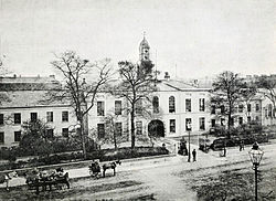 A black and white photograph, showing a stately-looking two-storey building with white walls, extending out of shot to the left and right, with an arched cart entrance at the centre. A modest clocktower rises above the entrance, and the building is surrounded by neat shrubbery and iron railings. A wide street crosses left-right outside of the fence, with a handful of horse-drawn carts and pedestrians in 19th century clothing.