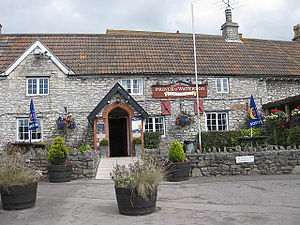 Stone building with tiled roof. Sign saying Prince of Waterloo. In the foreground are tables, benches and planters.